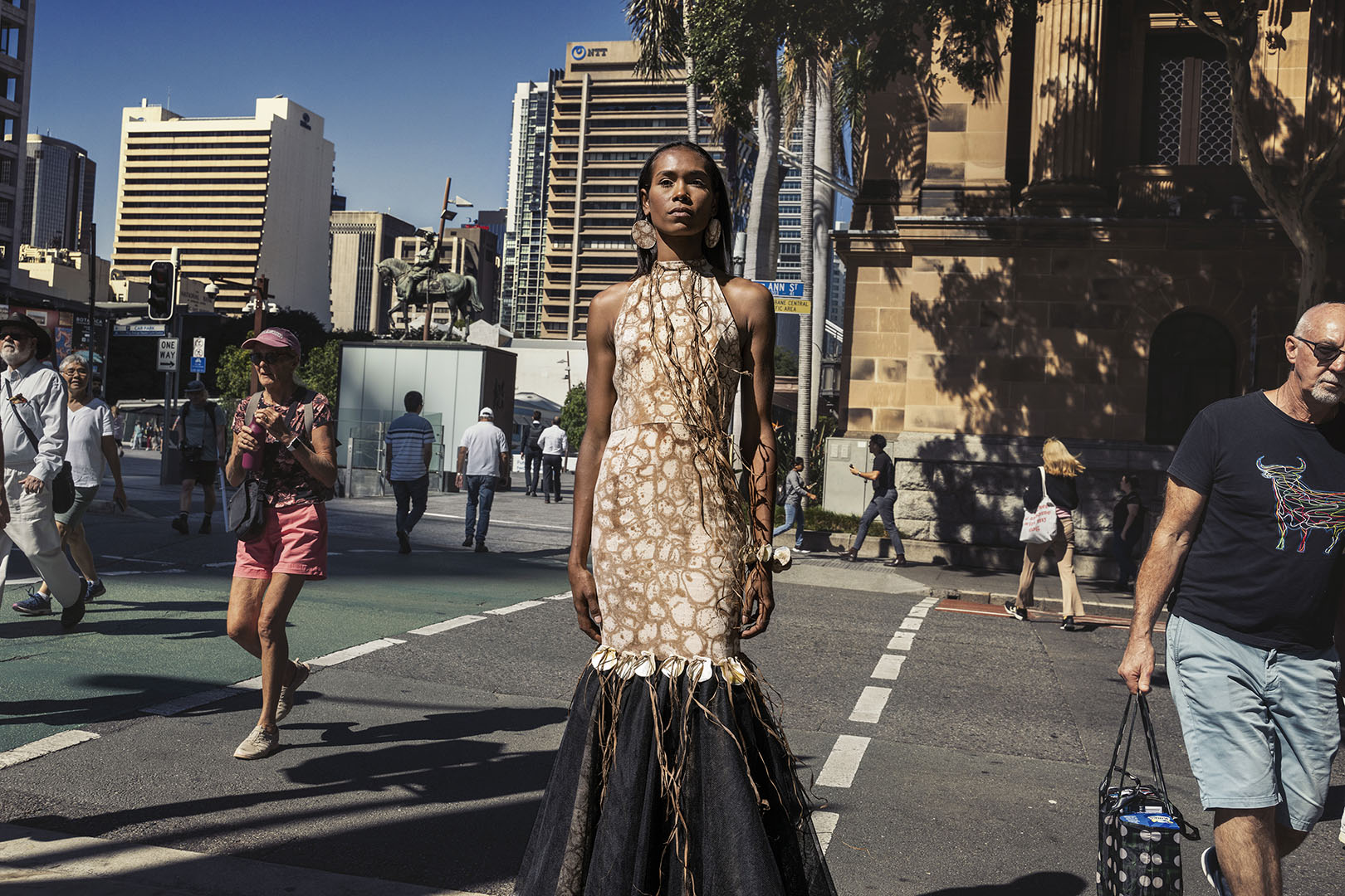 A striking fashion photograph featuring a model in an earth-toned gown standing confidently on a city crosswalk, with pedestrians and architecture in the background.