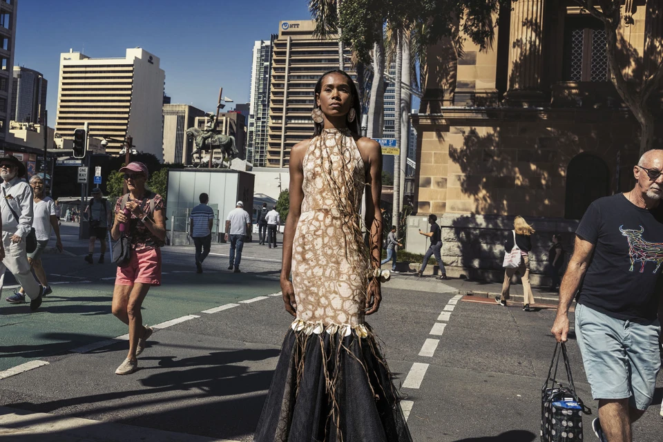 A striking fashion photograph featuring a model in an earth-toned gown standing confidently on a city crosswalk, with pedestrians and architecture in the background.