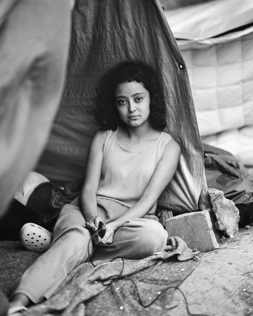 Young woman with curly hair sits inside tent, holding a cable. Black and white portrait shows relaxed facial expression.