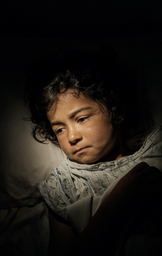 Portrait of a child with dark hair and eyes, looking down with a somber expression, against a dark, shadowy background in dim lighting.