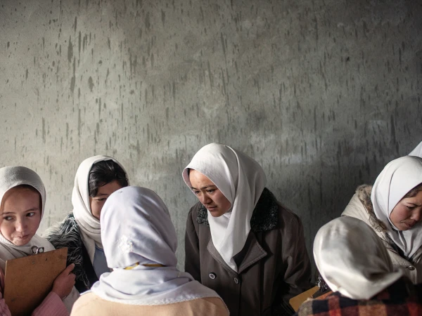 A group of Middle Eastern women in headscarves gather indoors, some holding clipboards, against a mottled gray background, indoors.
