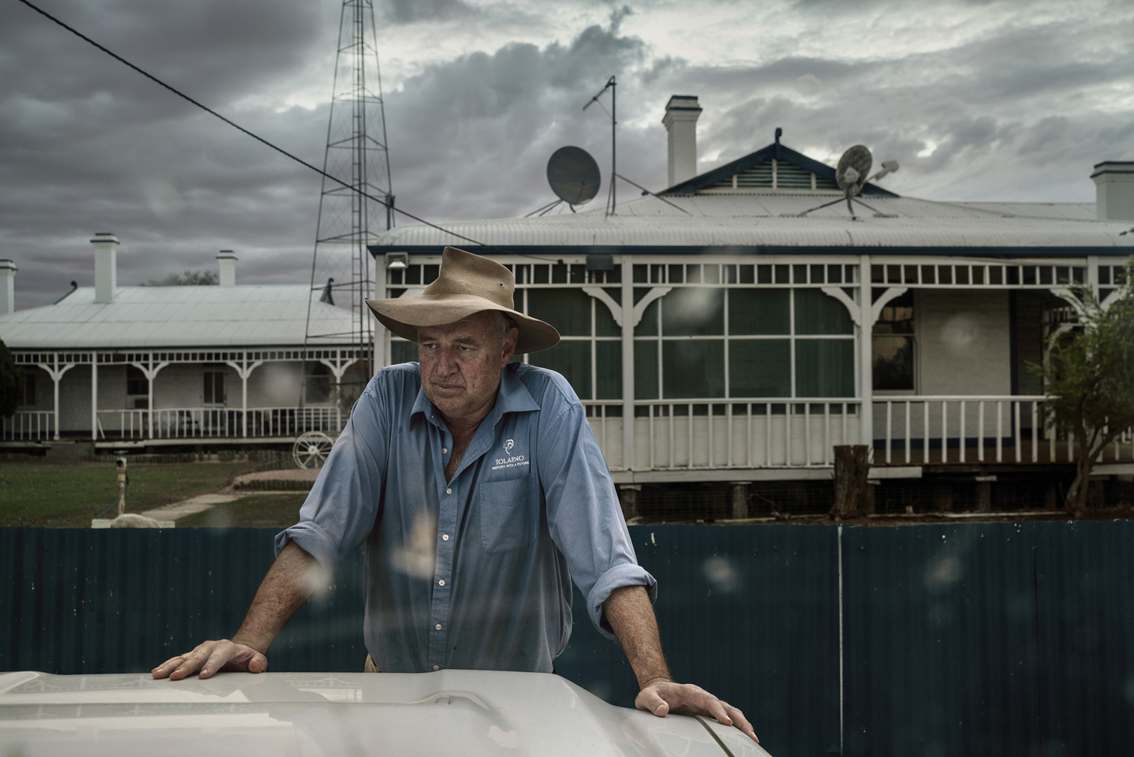 Man in hat and blue shirt with houses and antenna in background, outdoors on overcast day. Farmer standing outside, looking thoughtful.