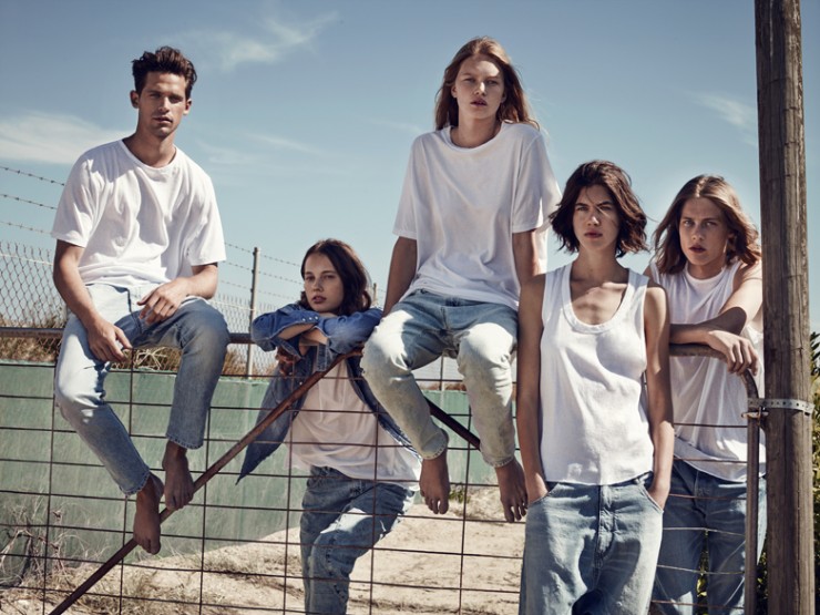 Five young adults pose outdoors in white tops and jeans, arranged on a metal fence against a bright blue sky and neutral rural background.