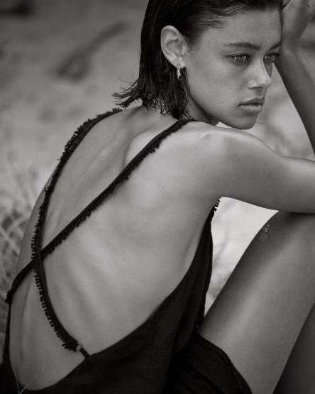 Monochrome portrait of a woman with wet, short hair, wearing a dark dress with straps, gazing to the right, set against a neutral background.