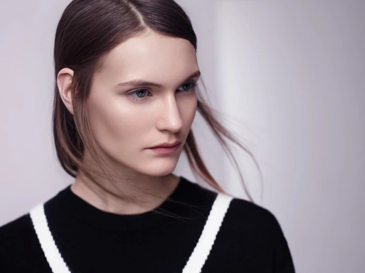 Close-up studio portrait of a young adult woman with fair skin, light eyes, and dark hair, wearing a black sweater with white stripes.