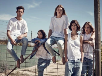 Group of five young models in casual white t-shirts and denim jeans posing outdoors against a wire fence under a clear sky.