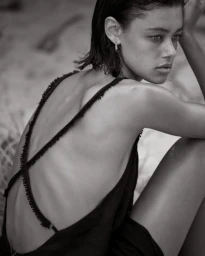 Black and white portrait of a woman with wet hair wearing a dark backless dress with thin cross straps, seated outdoors.