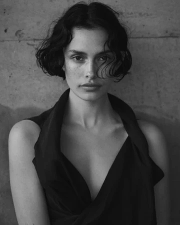 Monochrome portrait of a woman with short dark wavy hair wearing a sleeveless V-neck top against a textured concrete wall.