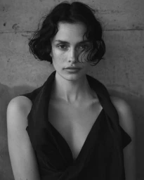Monochrome portrait of a woman with short dark wavy hair wearing a sleeveless V-neck top against a textured concrete wall.