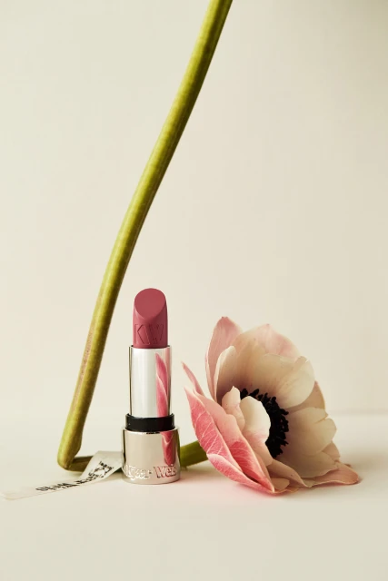 Close-up studio shot of a pink lipstick next to a pink and white anemone flower with a green stem on a light background.