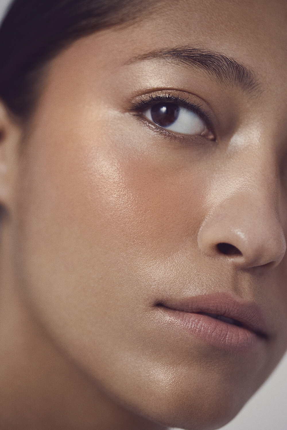 Close-up portrait of an adult female, showcasing her makeup and skincare. Her skin tone is bronze, with soft lighting, and neutral background.