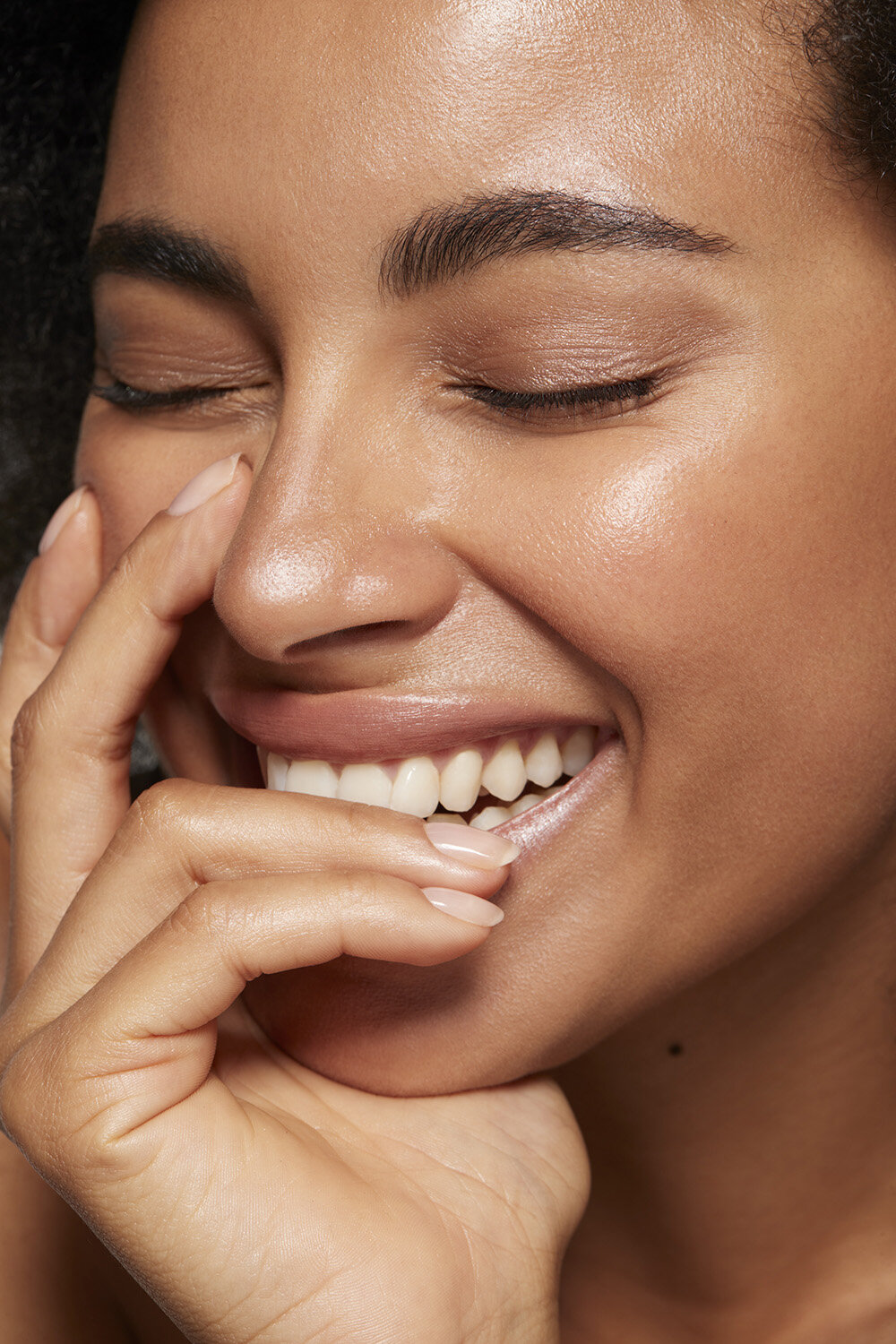 Close-up of a smiling adult female with dark hair and tanned skin, her hand near her face in a softly lit, indoor portrait.
