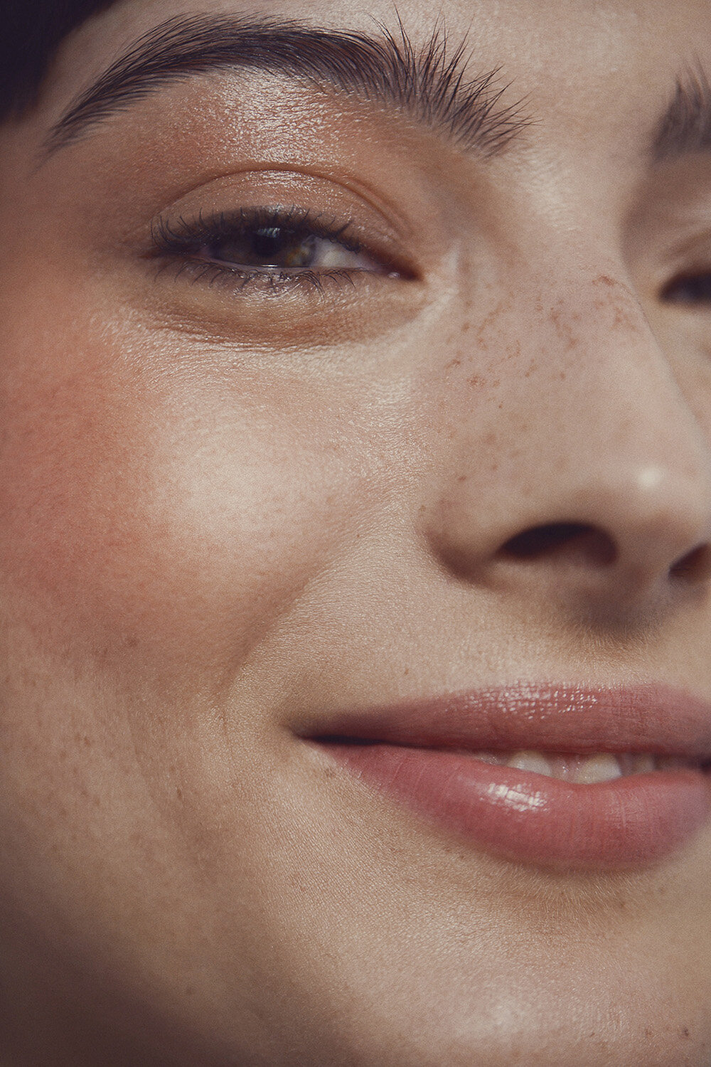 Close-up of a smiling young woman with fair skin and freckles, showcasing natural makeup with glossy lips and highlighting facial features.