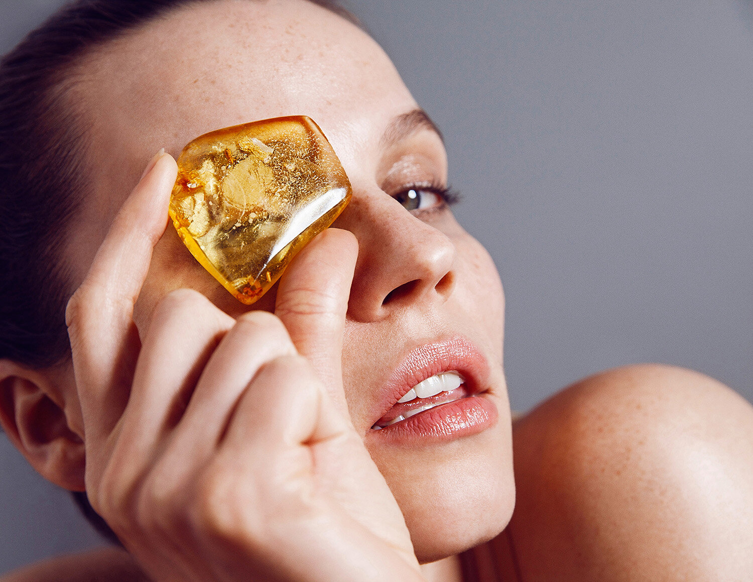 Close-up studio shot of a fair-skinned adult female holding a transparent yellow amber stone to her eye with a neutral facial expression.