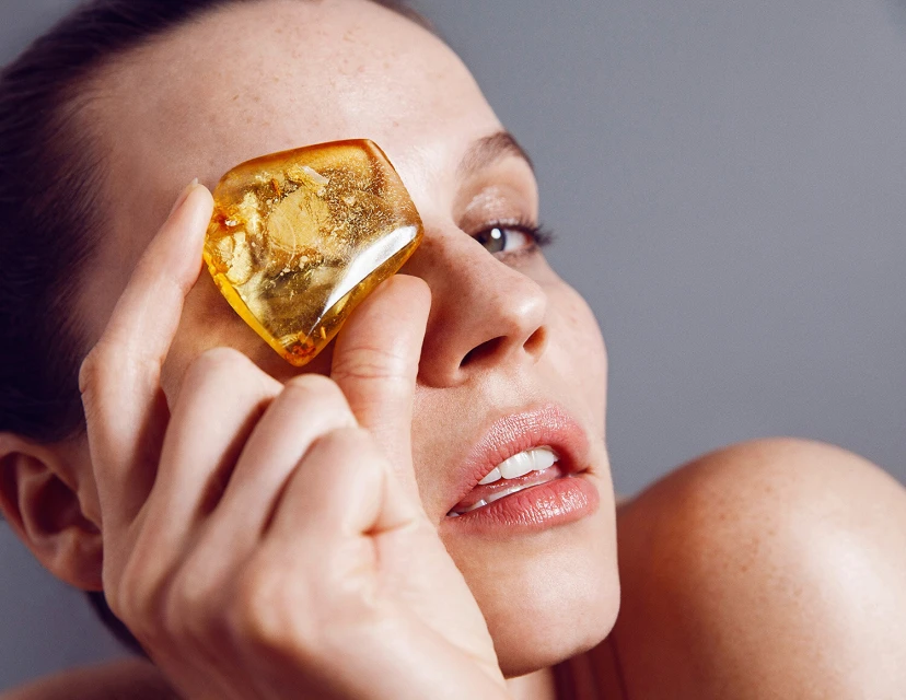 Close-up studio shot of a fair-skinned adult female holding a transparent yellow amber stone to her eye with a neutral facial expression.
