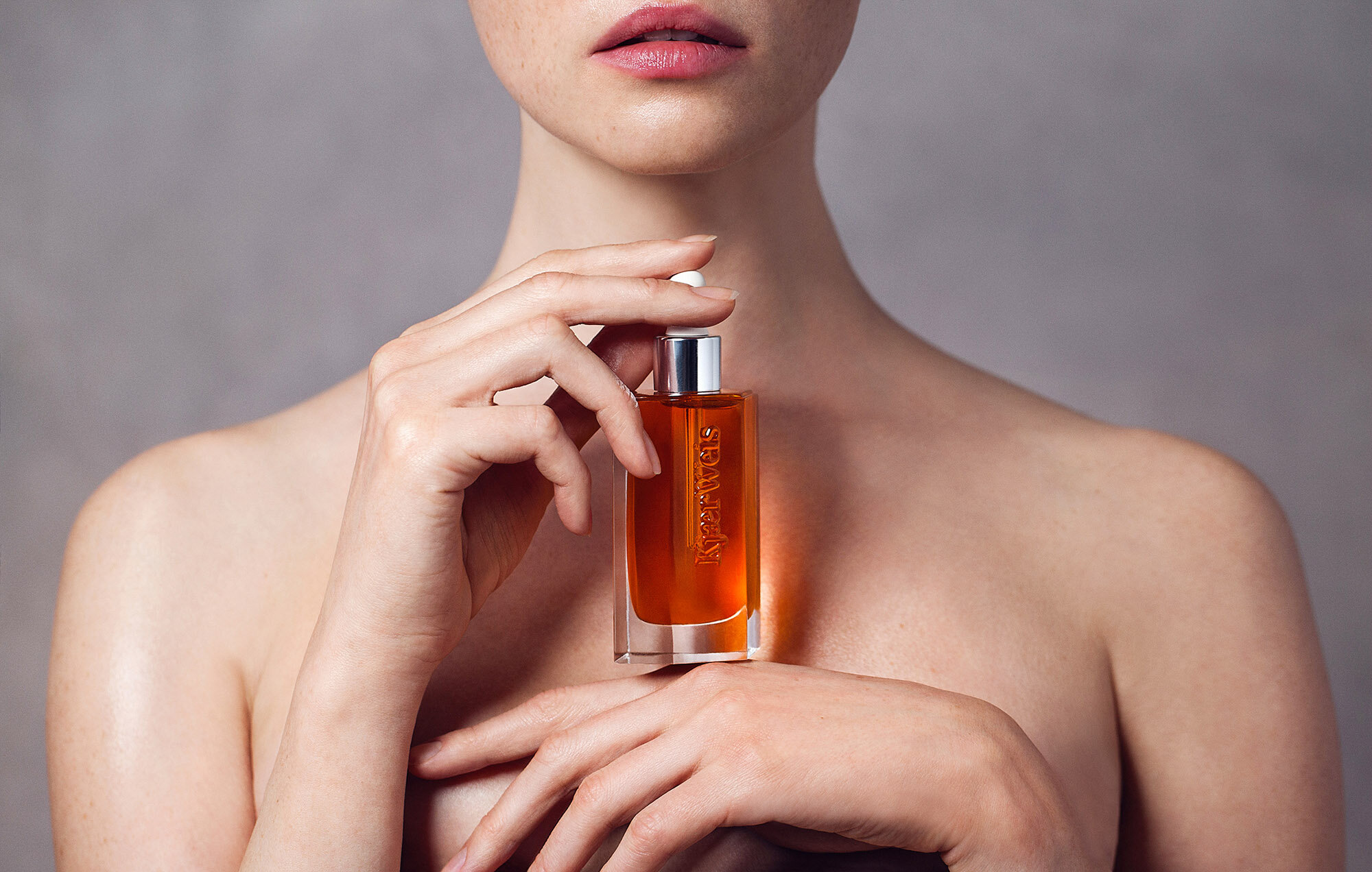 Elegant portrait of woman holding bottle of amber beauty oil; hand close-up, studio lighting, muted grey background. Beauty and skincare.