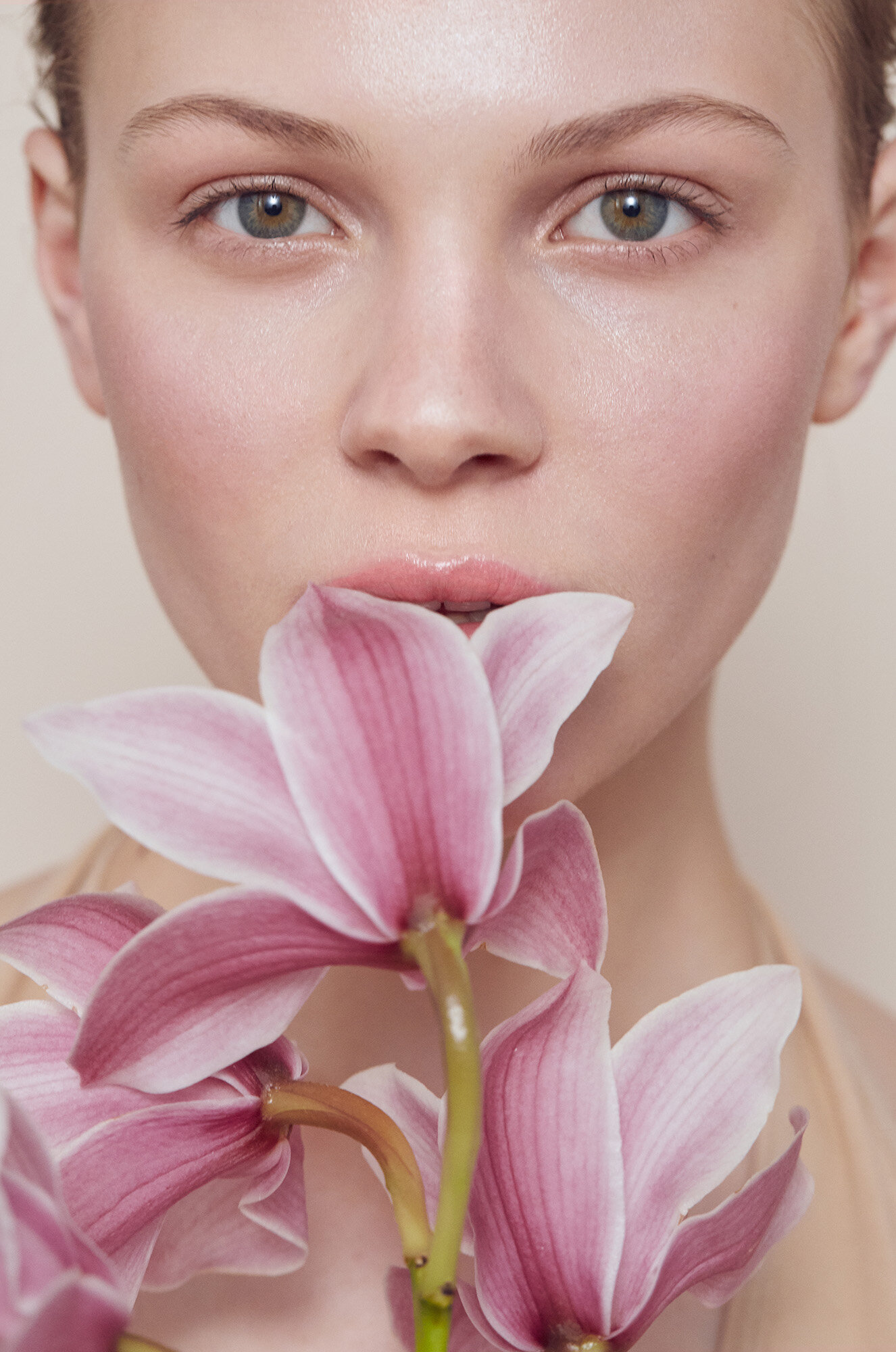 Close-up portrait of a fair-skinned young woman with pink orchids covering her mouth against a neutral backdrop, beauty shoot.
