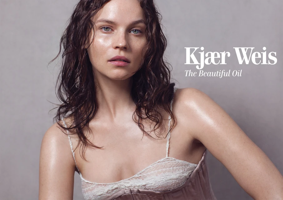Close-up beauty shot of an adult female model with wet dark hair and glossy skin wearing a light pink lace-trimmed camisole against a gray background.
