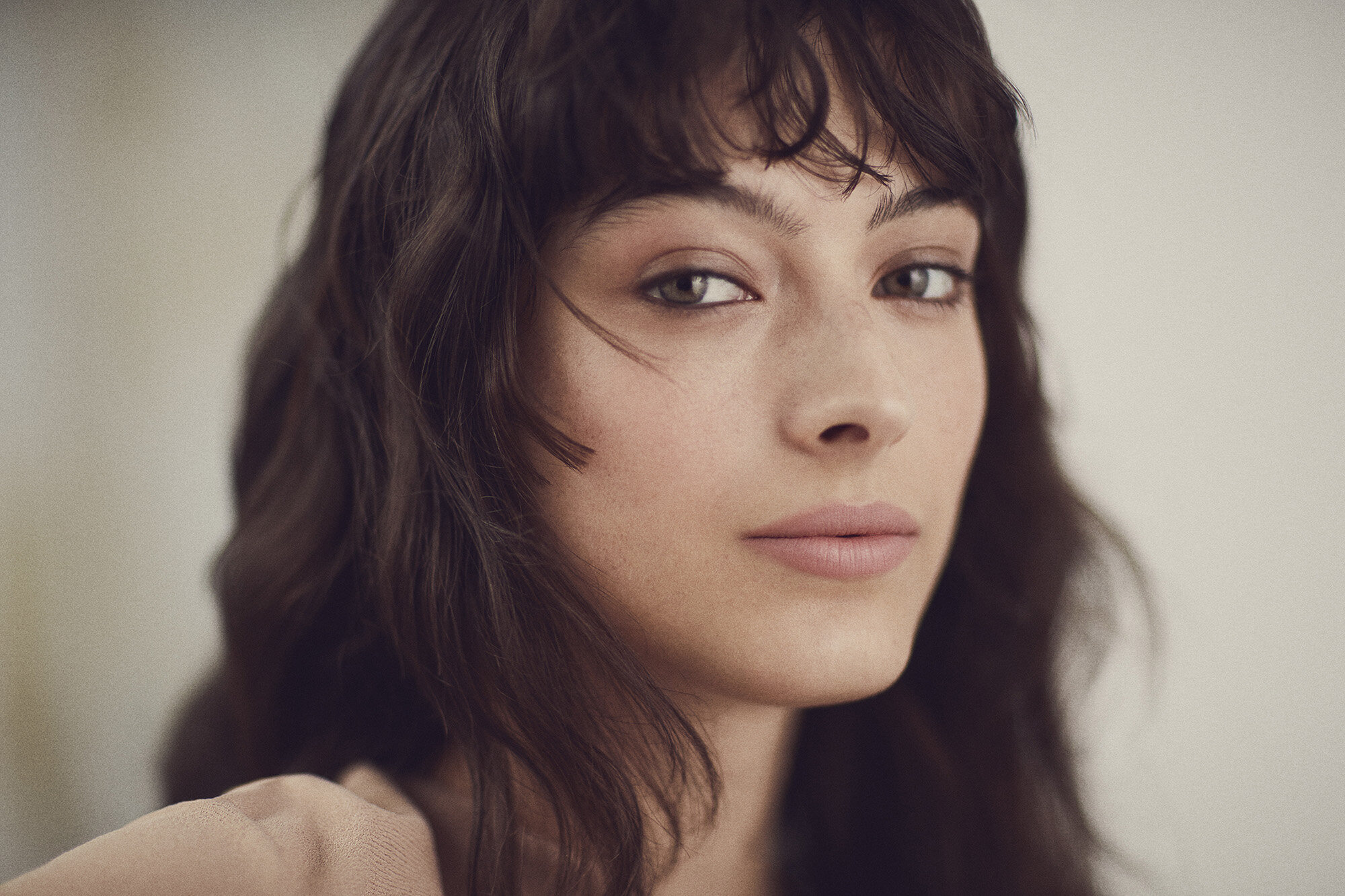 Close-up portrait of a young woman with wavy brunette hair and fair skin, gazing directly at the camera in a softly lit indoor setting.