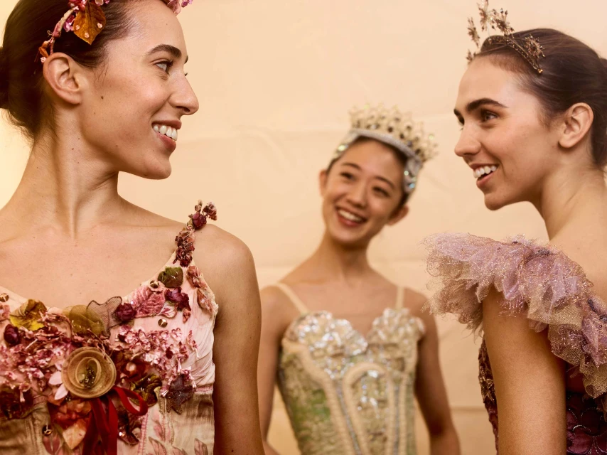 Three ballerinas in ornate costumes and floral headpieces smile in an indoor setting, showcasing intricate detailing and warm lighting.