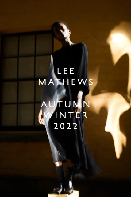Fashion shot of a Black model wearing a black dress, black socks and shoes, posing in front of a wall with window shadows and 'Lee Mathews' text.