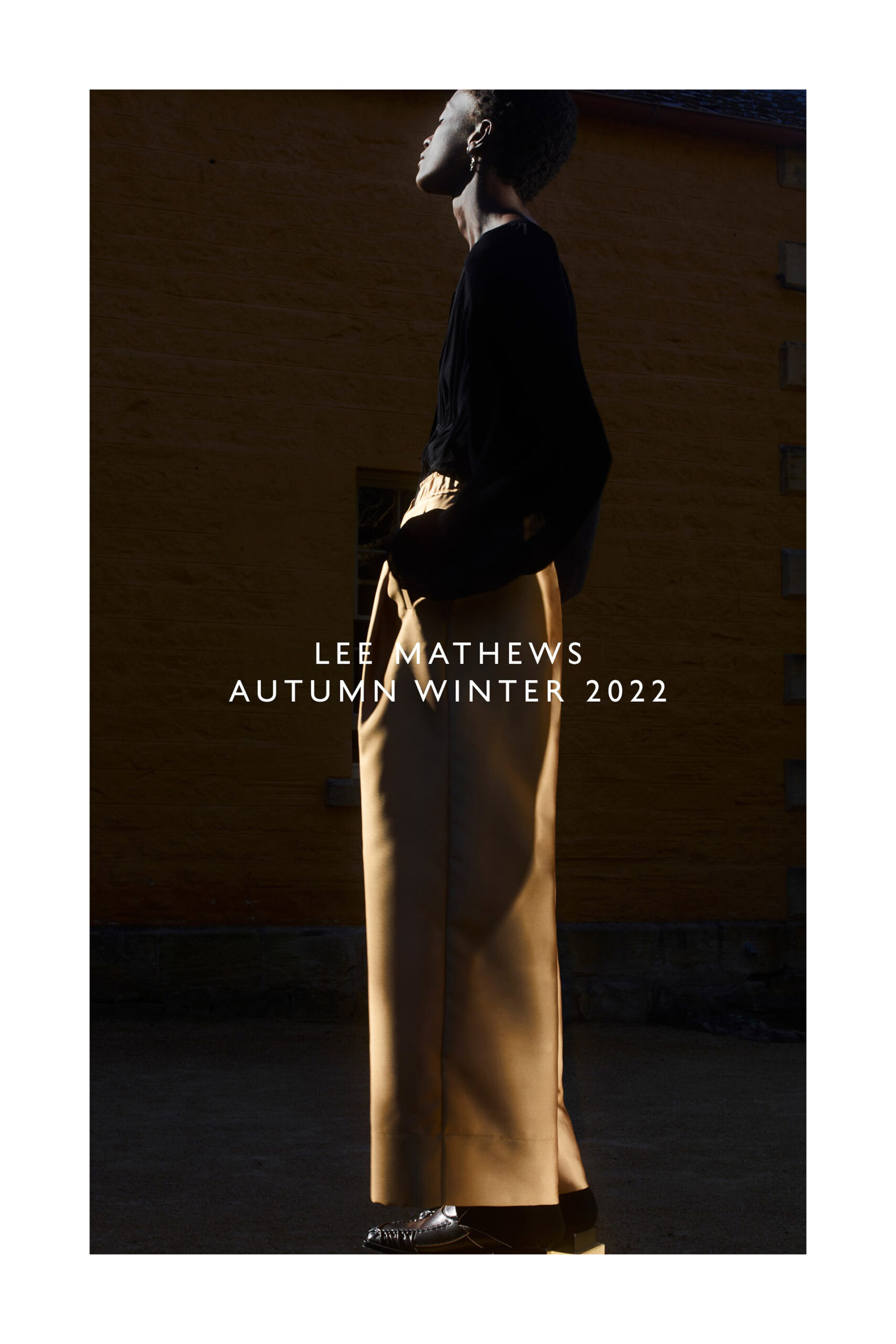 Fashion editorial shot of a slender, dark-skinned woman wearing a black top and tan pants, set against a textured wall.