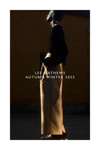 Fashion editorial shot of a slender, dark-skinned woman wearing a black top and tan pants, set against a textured wall.