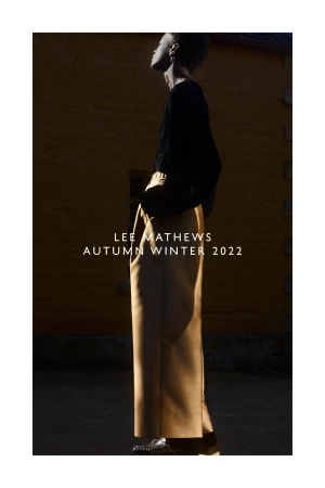 Fashion editorial shot of a slender, dark-skinned woman wearing a black top and tan pants, set against a textured wall.