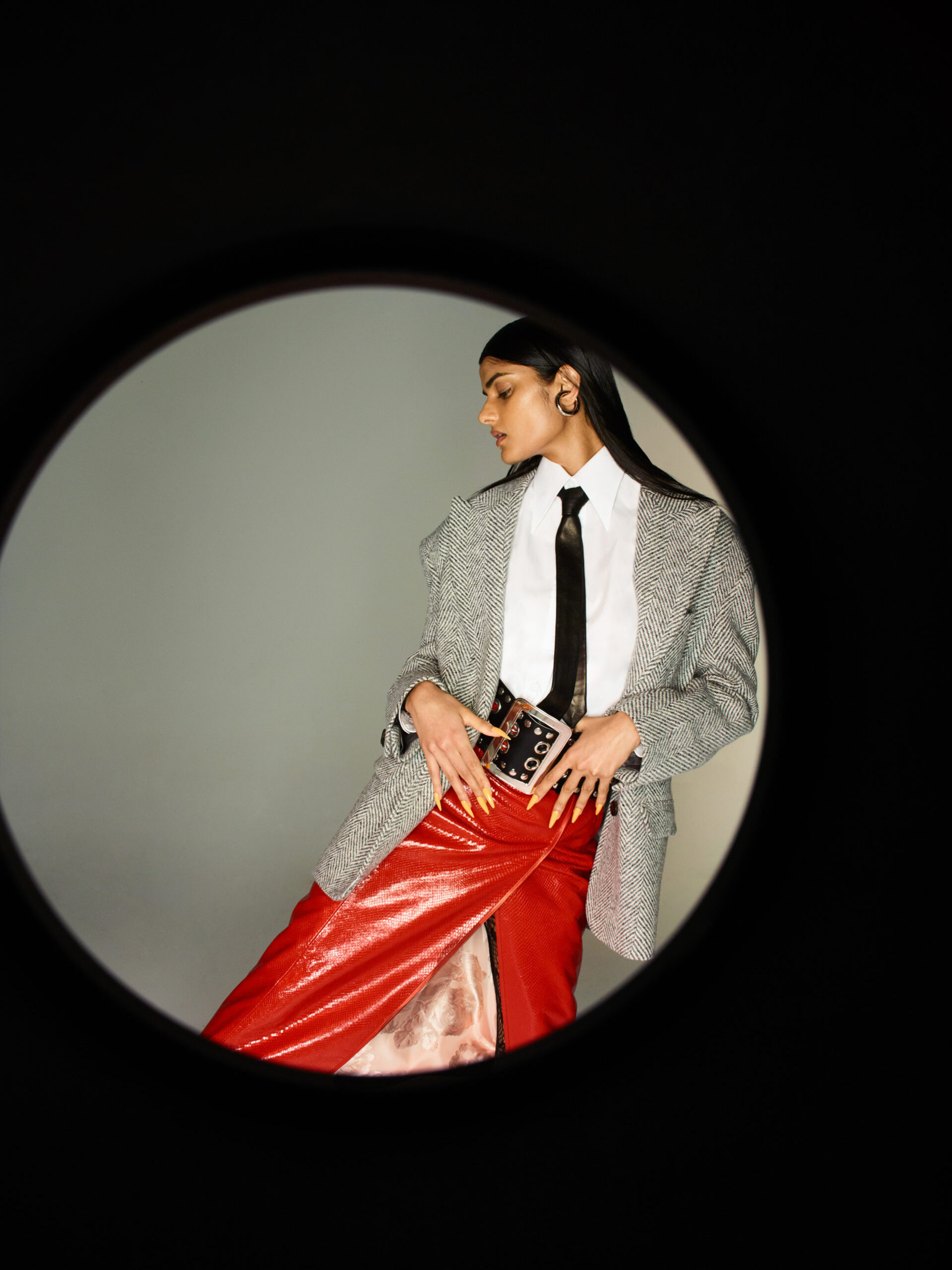 Fashionable woman poses in red pants, white button-down shirt, and grey jacket, framed by a black circular foreground element.