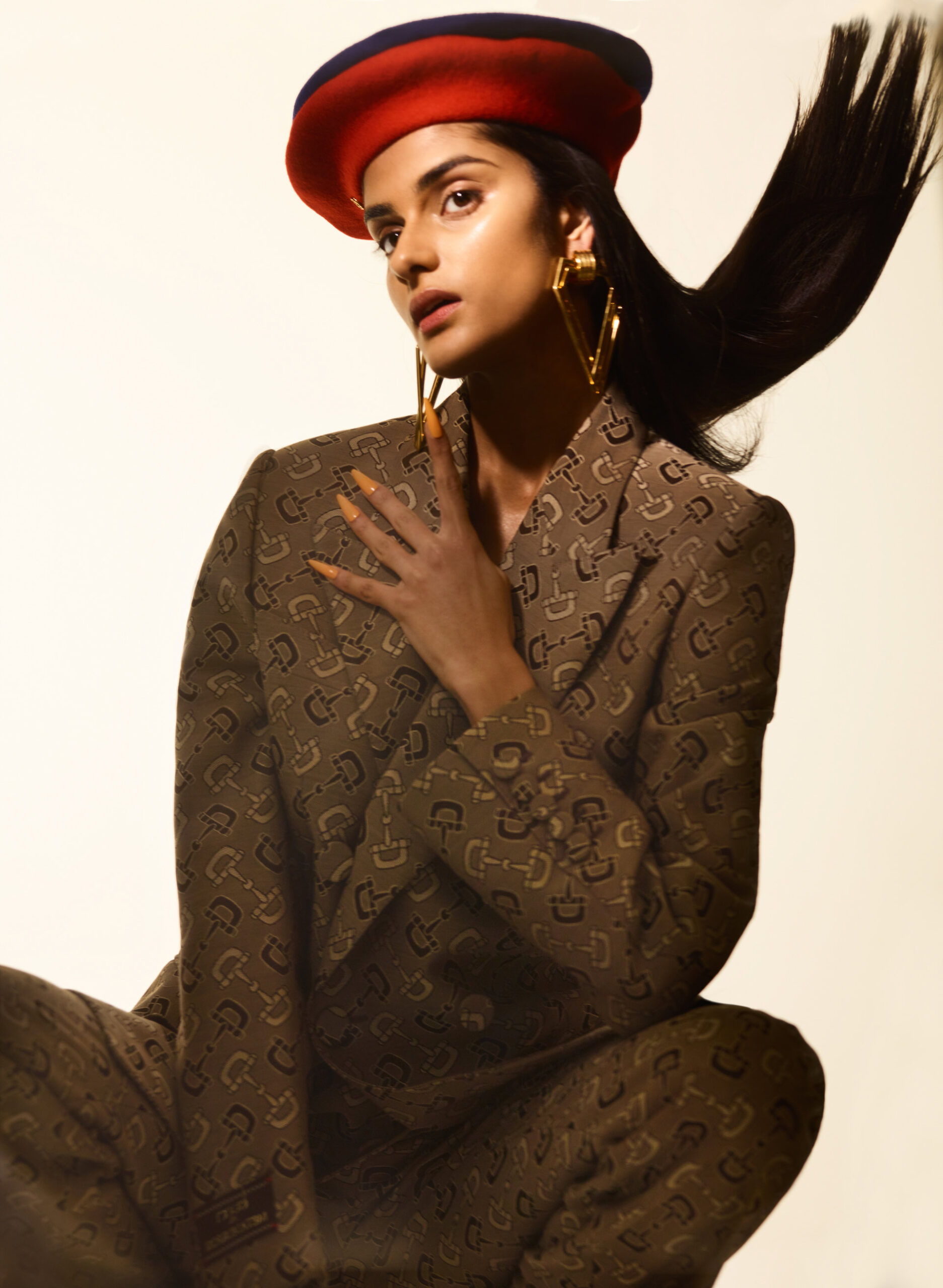 A woman in a patterned suit and red beret poses in a studio with one hand near her chest and hair flowing. She wears large gold earrings.