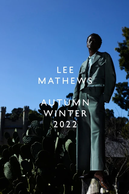 Fashion shot of a Black male model outdoors, posing in a sage-green suit with cactus plants, a building, and a blue sky in the background.