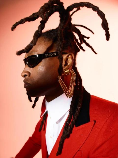 Portrait of a Black man with dreadlocks wearing a red suit, white shirt, large gold earrings, and Gucci sunglasses against a red backdrop.