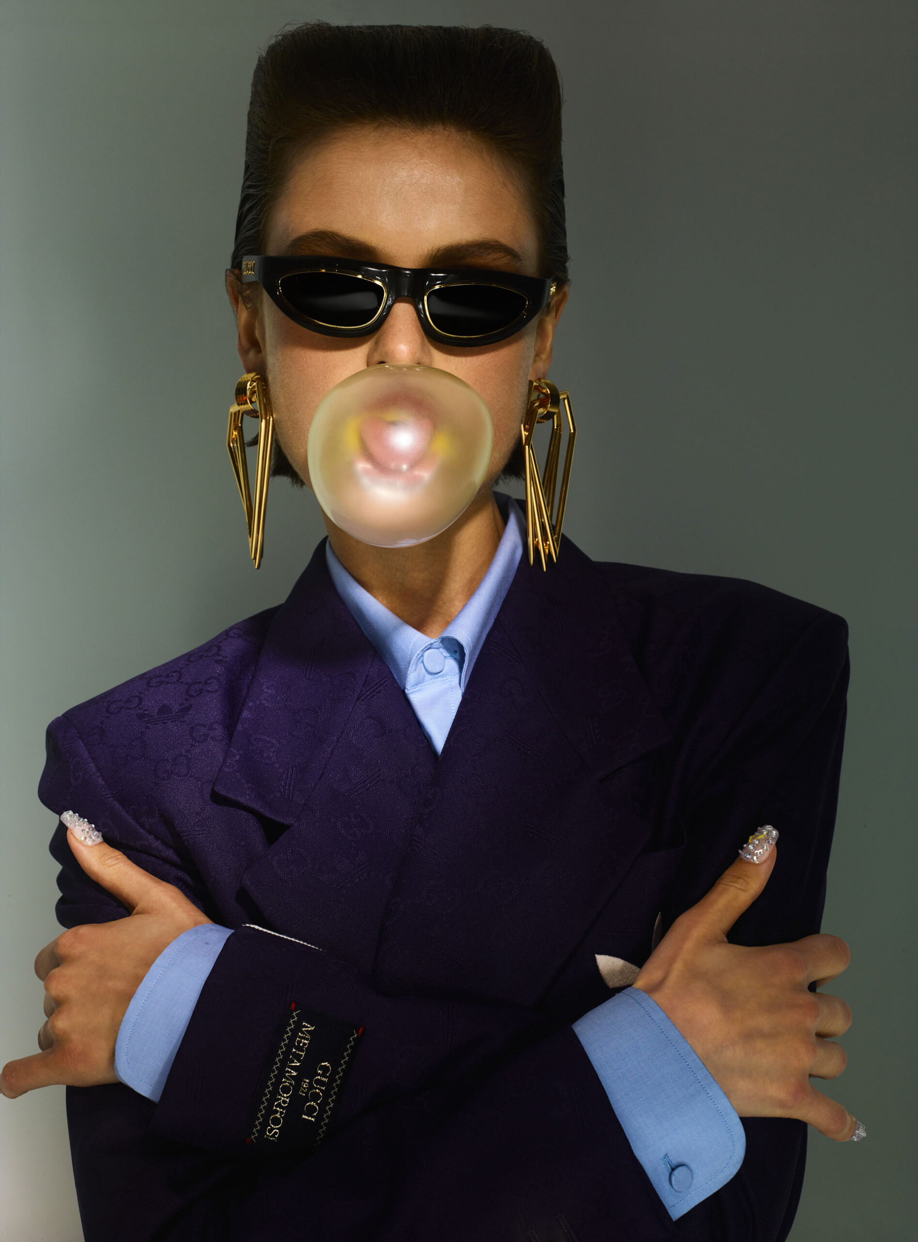Stylish woman with bubblegum bubble, wearing dark blazer, light blue shirt, sunglasses, and long gold earrings against grey background.