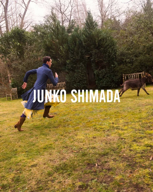 A person in a blue coat runs across a grassy lawn towards a donkey in an outdoor setting, trees and greenery in the background.