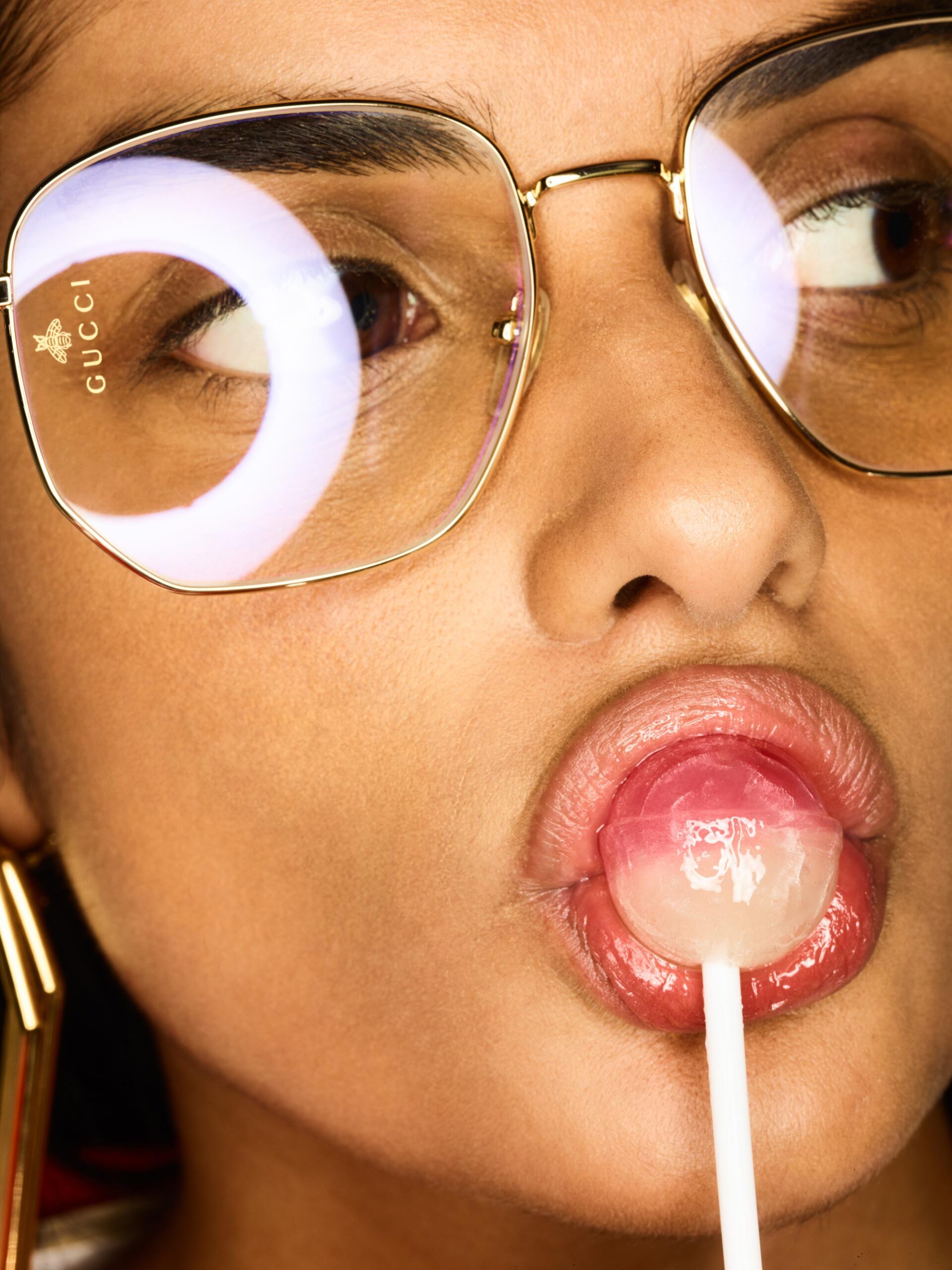 Close-up of a woman with tan skin eating a lollipop, wearing gold glasses and gold earrings with red lipstick against a warm background.