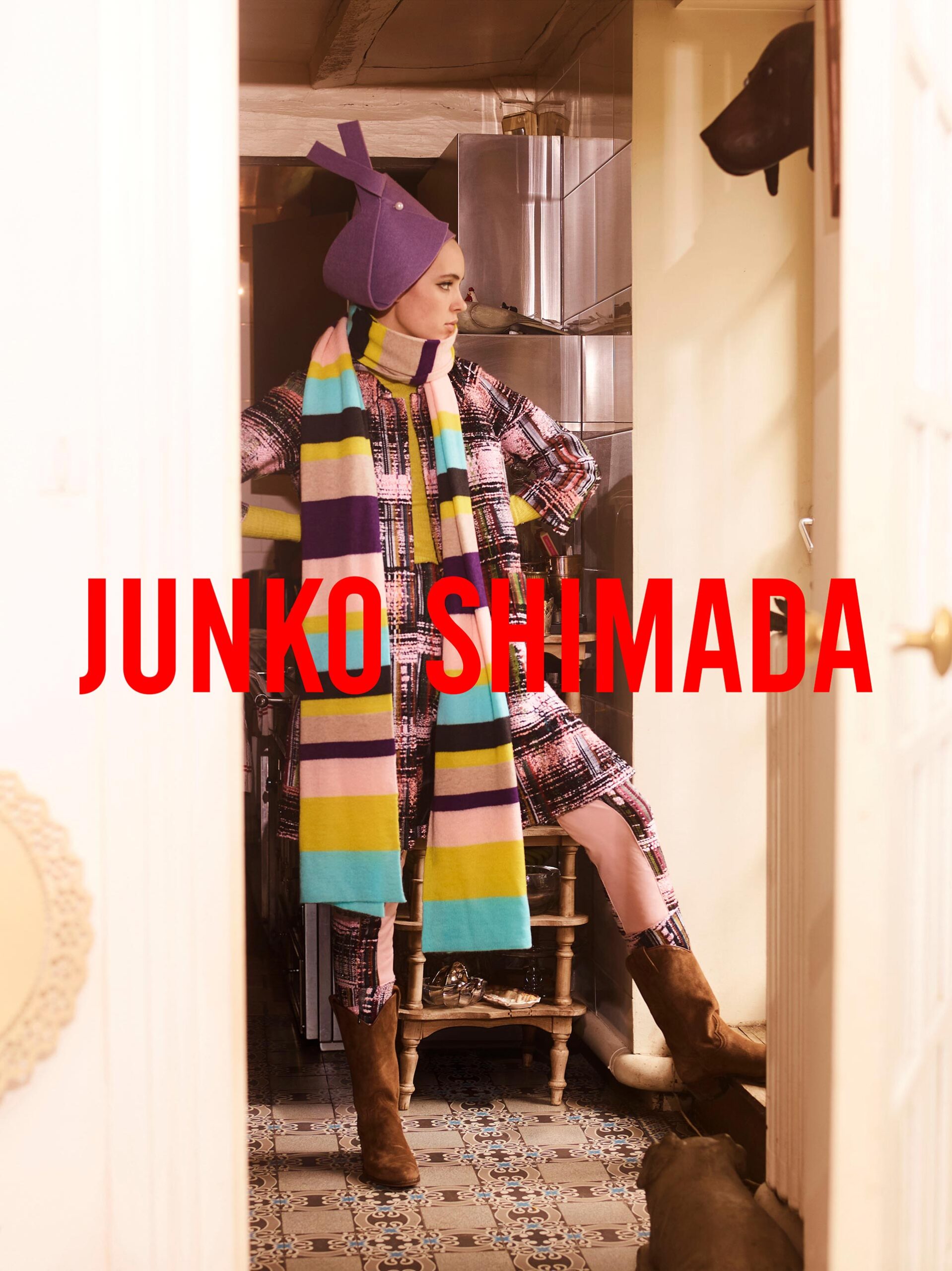 A woman in a patterned coat, hat, and scarf stands in a doorway, showcasing a fashion-forward ensemble with unique accessories.
