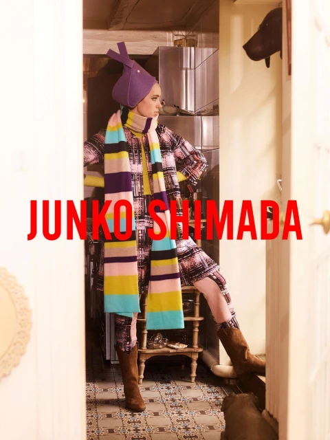 A woman in a patterned coat, hat, and scarf stands in a doorway, showcasing a fashion-forward ensemble with unique accessories.
