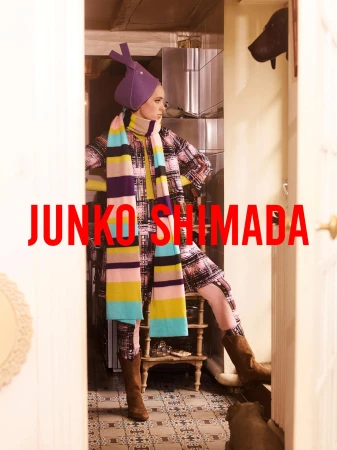 A woman in a patterned coat, hat, and scarf stands in a doorway, showcasing a fashion-forward ensemble with unique accessories.