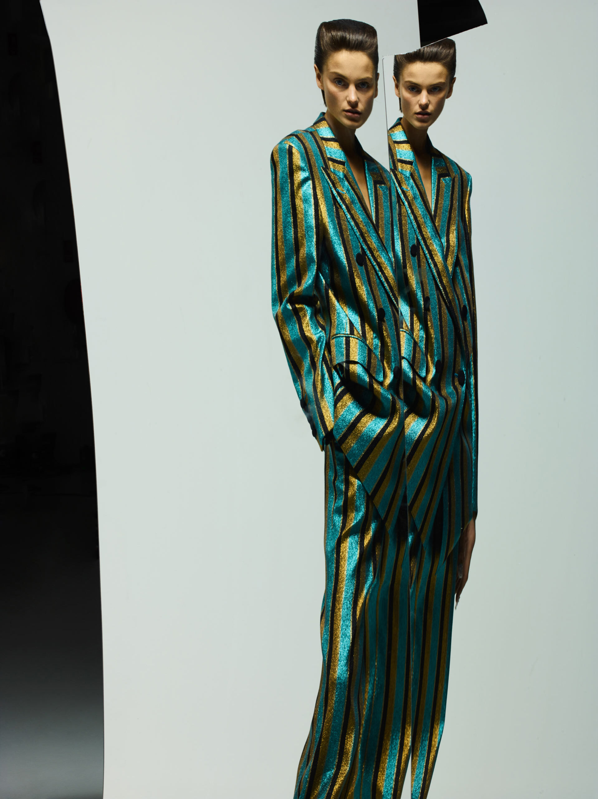 Two women pose indoors wearing striped teal and gold suits in a fashion editorial style against a white background with a black object.