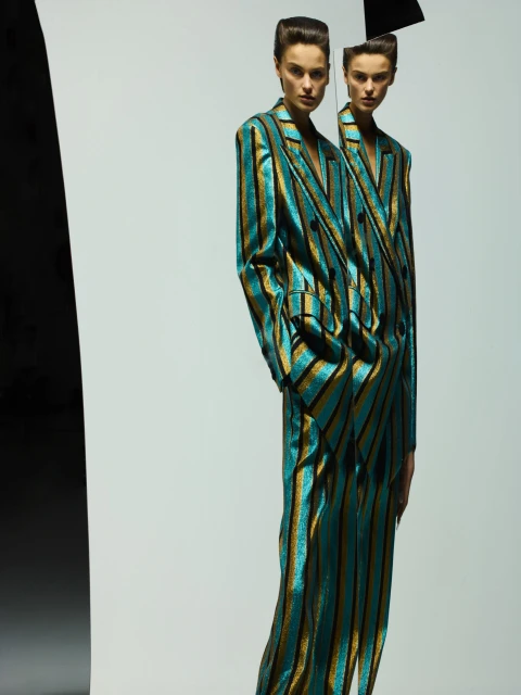 Two women pose indoors wearing striped teal and gold suits in a fashion editorial style against a white background with a black object.