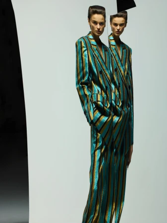 Two women pose indoors wearing striped teal and gold suits in a fashion editorial style against a white background with a black object.