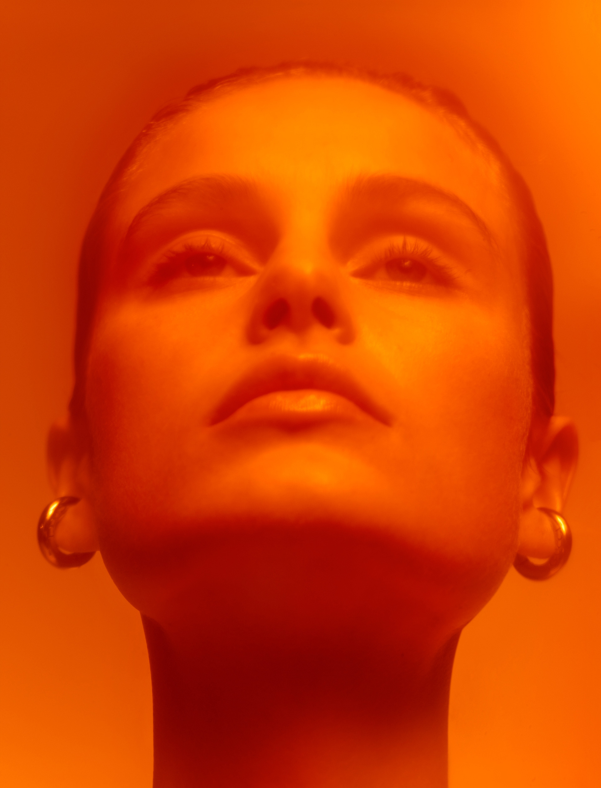 Close-up portrait of a young woman with her head tilted back looking up, wearing gold hoop earrings, bathed in orange lighting.