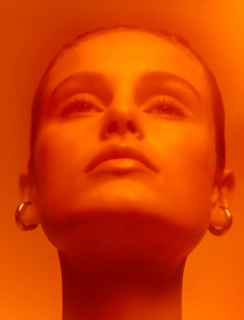 Close-up portrait of a young woman with her head tilted back looking up, wearing gold hoop earrings, bathed in orange lighting.