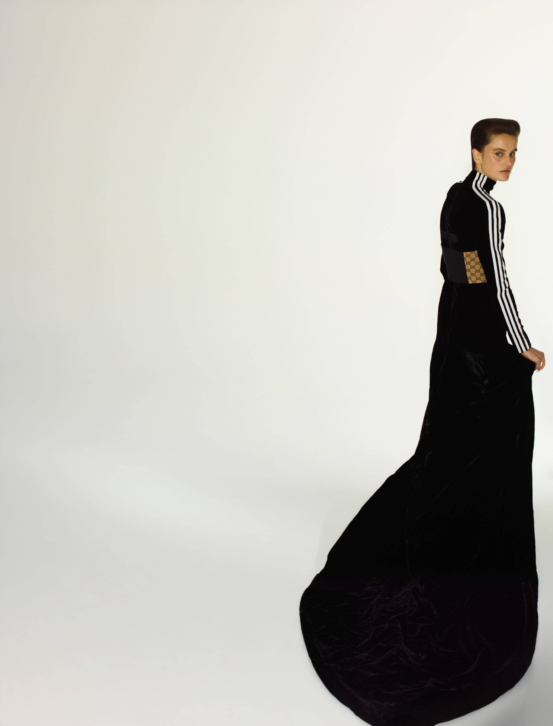 Woman in long black gown with Adidas stripes posing in a brightly lit studio. Fashion photography with a full-length portrait on white.