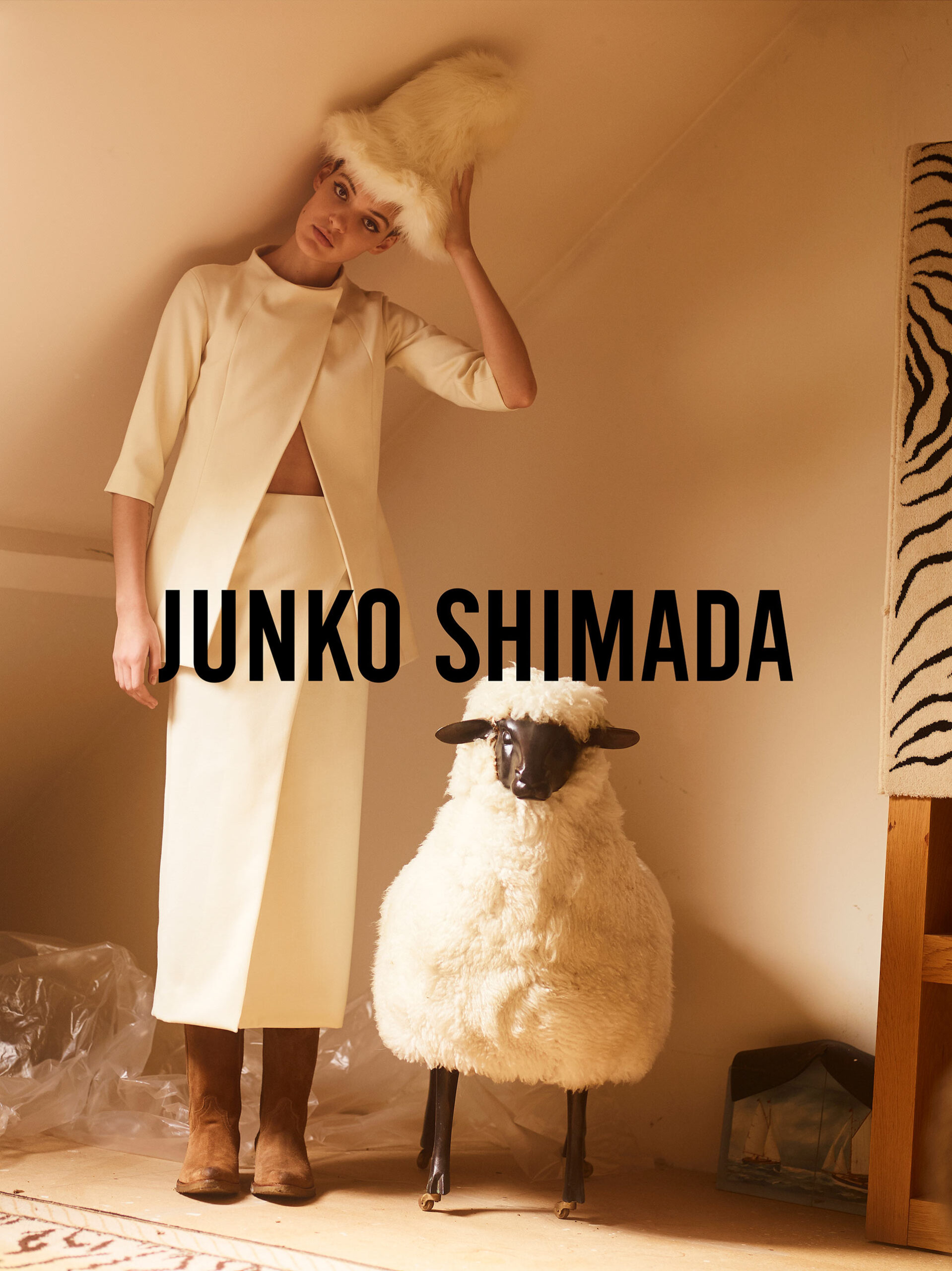Woman in cream skirt-suit and furry hat poses next to a stuffed sheep in a room with natural light for a fashion editorial.