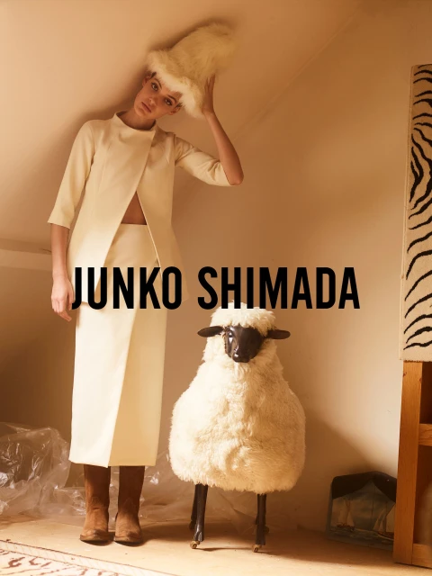 Woman in cream skirt-suit and furry hat poses next to a stuffed sheep in a room with natural light for a fashion editorial.