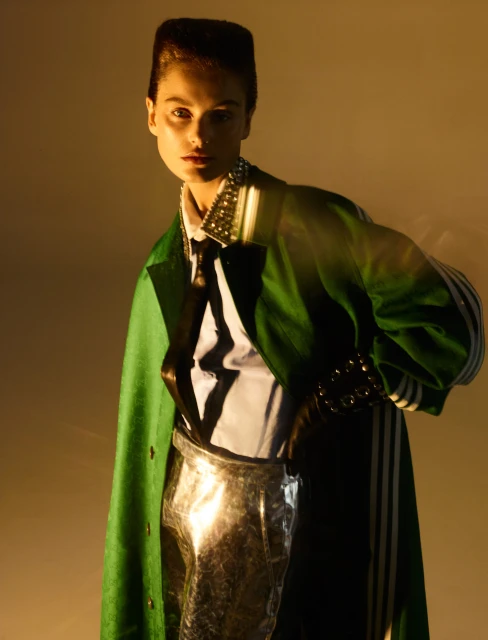 Fashion portrait of a woman with dramatic makeup wearing a green coat with metallic silver pants in a studio with amber lighting.