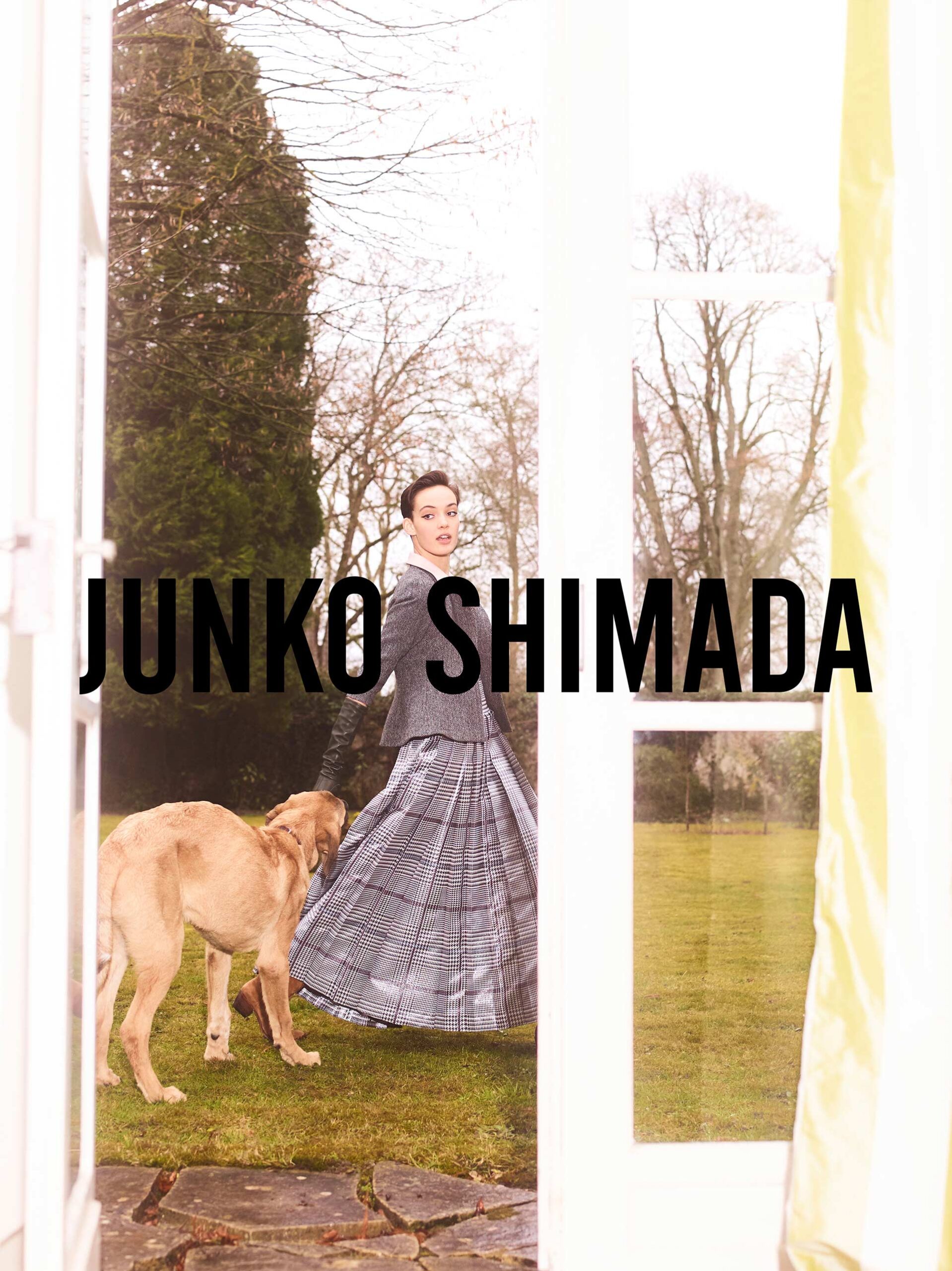 Fashion shot of a woman in a grey skirt and jacket, walking with a golden retriever outdoors, seen through a doorway, plus brand text.
