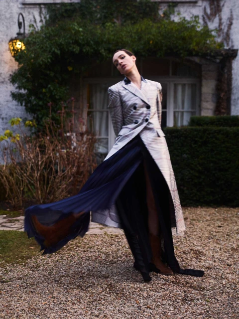 Elegant woman in a grey blazer and long blue skirt poses in front of a vine-covered building, creating a sophisticated fashion moment.