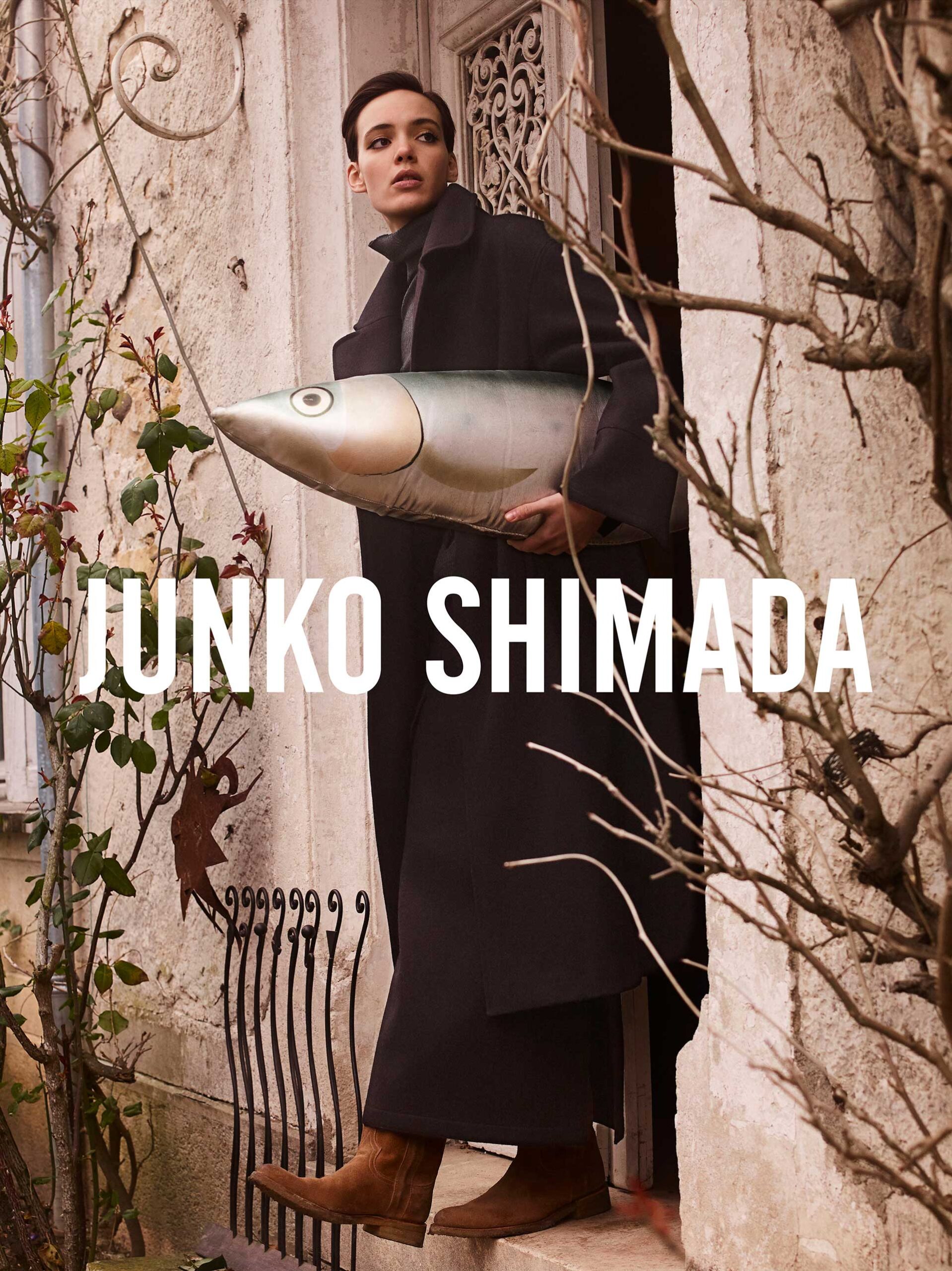 A woman in a black coat holds a large fish-shaped object against a textured wall with climbing plants and the words JUNKO SHIMADA.
